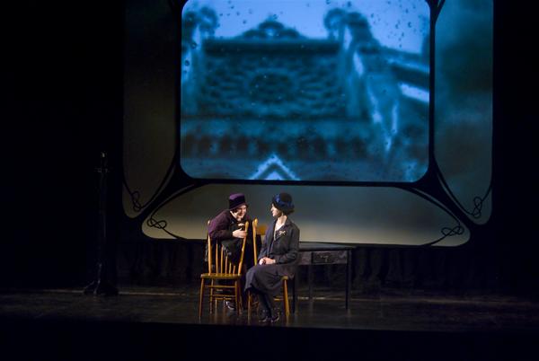 Two actors are performing on a dimly lit stage with wooden chairs and a table. A black-and-white image is projected behind them, showing an ornate building. Both are dressed in dark, vintage-style costumes.