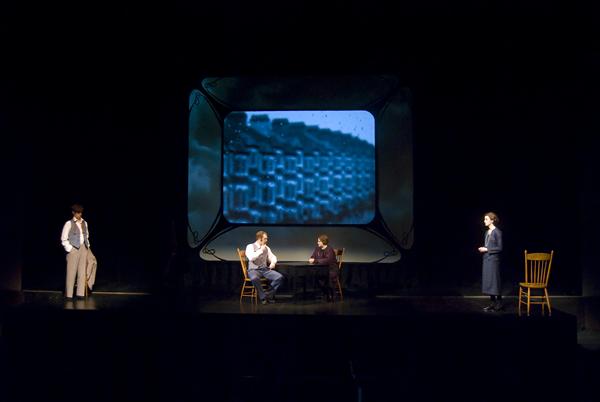 Four actors are on a dimly lit stage during a theatrical performance. A large screen displays an abstract pattern in the background. Two actors sit at a table, while the others stand separately, with two wooden chairs onstage.