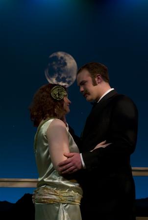 A couple gazes at each other under a full moonlit sky. The woman wears a cream dress with a floral headband, and the man is in a black suit. They are standing on a balcony with a mountainous backdrop and night sky.