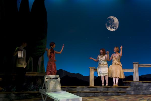 Four performers are on stage dressed in period costumes. Two in gold dresses are gesturing on the right, one in a red outfit stands in the center, and another with a lantern is partially visible on the left. A moonlit sky forms the backdrop.