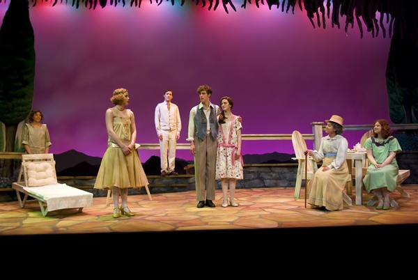 A group of actors on stage, performing in a play set outdoors. They are dressed in vintage clothing, standing on a stone-like floor with colorful lights above. Trees and a lounge chair are part of the set design. The background is a soft, gradient purple.