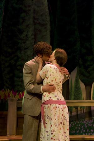 A couple shares a kiss on stage, surrounded by a garden backdrop with tall trees and flowers. The man is wearing a suit, while the woman is in a floral dress with a pink sash.
