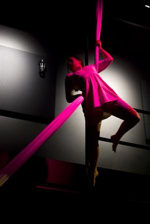 A person performs aerial silk acrobatics under dim lighting, highlighted with pink and red tones. They are suspended gracefully, with one leg bent and holding onto the silk fabric. The background is dark, enhancing the dramatic effect.