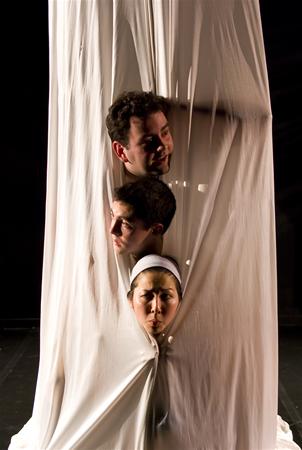 Three people press their heads through a tight, white fabric, creating distorted impressions on the material. The scene is dimly lit against a dark background, highlighting the unusual and creative concept.