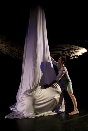 A performer on stage interacts with a tall, flowing piece of fabric under dramatic lighting. The fabric cascades from above, and the stage is dimly lit, creating a striking, shadowy effect. The performer appears focused and poised.
