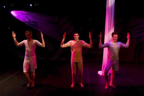 Three performers on stage under colorful lights, each with arms raised. They are dressed in form-fitting attire, and there is a fabric drape hanging in the background. The scene has a dynamic, theatrical atmosphere.