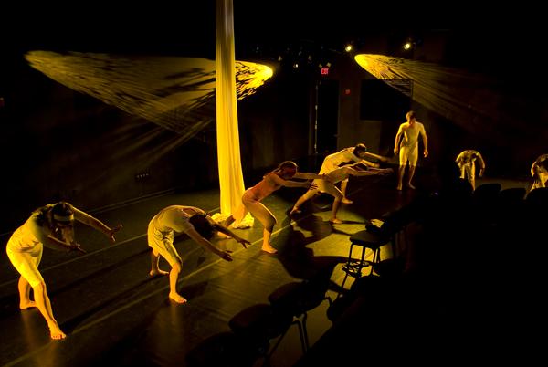 A group of dancers in white costumes perform expressive movements on a dimly lit stage. A spotlight creates dramatic shadows, and a white fabric hangs from the ceiling, enhancing the artistic atmosphere.