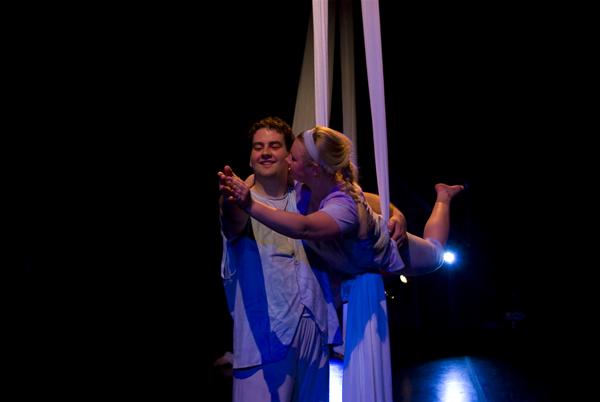 A man and a woman perform an aerial dance on white silks. The woman, suspended, kisses the man's cheek as he stands on the ground, smiling with eyes closed. They are on a dark stage with a spotlight in the background.