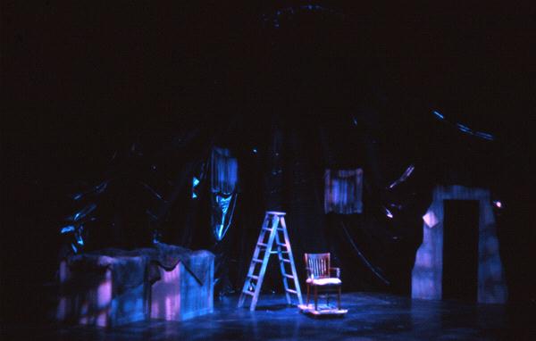A dimly lit stage features a wooden chair and a ladder. The backdrop is draped with dark, glossy fabric, creating a mysterious atmosphere. Subtle colored lights cast purple and blue hues onto the set.