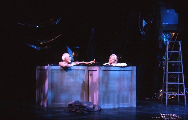 Two actors with silver hair are on stage, sitting in large bins, seemingly performing a dramatic scene. One reaches toward the other. The background is dark, with dim lighting highlighting parts of the set, including a ladder on the right.