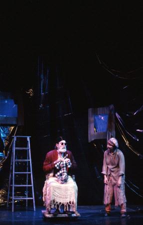 Two actors perform on a dimly lit stage. One is sitting in a wheelchair wearing a checkered blanket and dark glasses, the other stands nearby in ragged clothing. A ladder is to the left, and the backdrop has dark, shiny sheets with window cutouts.