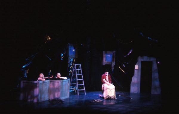 A dimly lit stage scene with three performers. Two are in a raised area on the left, and one is seated on a chair to the right. A ladder and dark drapes in the background create a moody atmosphere.