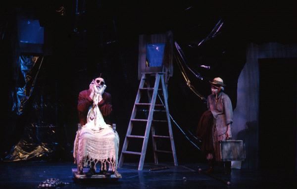 A dimly lit stage shows a person sitting in a chair covered with a tattered blanket, wearing dark glasses. Another person stands nearby holding a suitcase. A ladder and dark fabric hang in the background, creating a mysterious atmosphere.