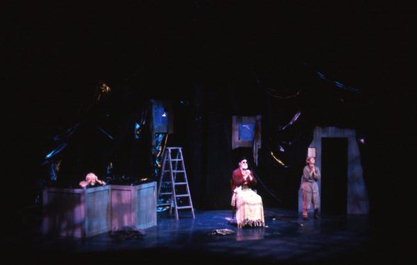 A dimly lit stage with a person sitting at a table in the center, surrounded by dark, abstract set pieces. A ladder is positioned to the left, and two people are near the table. The scene has a dramatic and mysterious atmosphere.