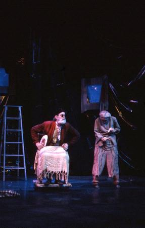 Two actors are on a dimly lit stage. One is seated with a blanket on the lap, wearing a dark jacket and hat. The other stands in ragged clothing, hunched over. A ladder and dark backdrop are visible.