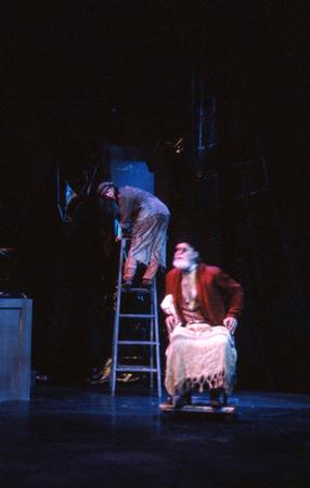 Two actors perform on stage in dim lighting. One is seated wearing a red jacket and knit blanket over their lap, while the other stands on a ladder in the background. The scene is set against a dark backdrop, evoking a dramatic atmosphere.