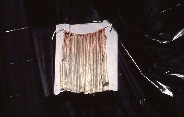 A light brown, pleated skirt hangs on a wall-mounted hanger against a dark, textured background. The fabric is illuminated, contrasting with the shadowy backdrop.