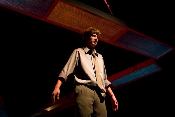 A man stands alone on a dimly lit stage, wearing a loose shirt and tie, with a spotlight shining from above. The background shows abstract geometric shapes, enhancing the dramatic atmosphere.