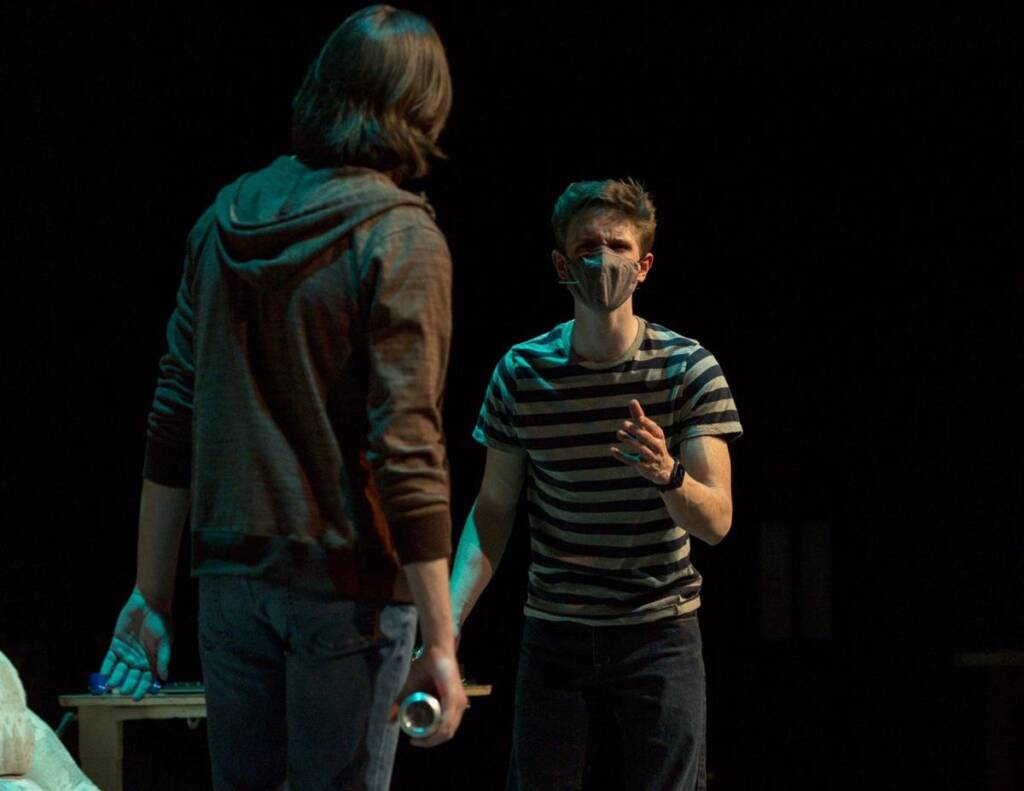 Two individuals are on stage, engaged in a dramatic scene. One person, wearing a striped shirt and mask, gestures expressively, while the other, in a hoodie, has their back to the camera. The lighting is subdued, creating a tense atmosphere.