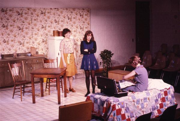 Three people are on stage in a room with a floral wallpaper. Two stand beside a kitchen area with a table and chairs, while the third sits on a bed with a patchwork quilt, looking into an open suitcase.