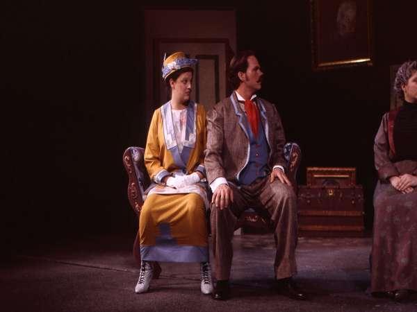 A man and a woman in early 20th-century attire sit side by side on a stage. The woman wears a yellow and blue dress with a matching hat, and the man is in a brown suit with a blue vest and red tie. They both face left, looking away.