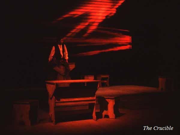 Stage set for "The Crucible" in dark, moody lighting with red and orange hues. A figure stands near a wooden table and benches. Dramatic shadows create an intense atmosphere, suggesting tension and drama.