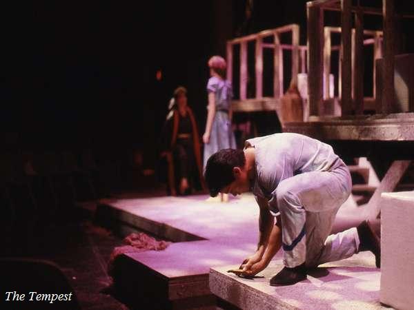 A person in work attire kneels and appears to work on the edge of a stage with a set depicting a wooden structure. Two other people stand in the background, one dressed in a light-colored dress. The setting is dimly lit with theatrical lighting.