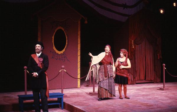 Three actors performing on a stage set resembling a circus tent. One actor stands apart, dressed in a dark outfit with a sash, while the other two, in colorful costumes, engage in animated gestures. A roped-off area and painted backdrop are visible.