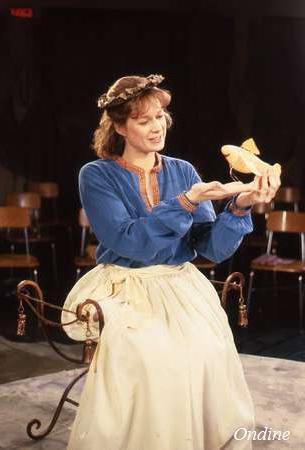 A person in a blue blouse and white skirt sits on a decorative chair, smiling at a puppet of a fish they hold in their hands. They wear a floral headpiece. Several wooden chairs are visible in the background. The scene is indoors.
