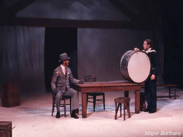 Two actors in a theater scene: one sitting at a wooden table dressed in a gray suit and top hat, and the other standing, holding a large drum. The background is minimal with a spotlight on the actors. The bottom right reads "Major Barbara.