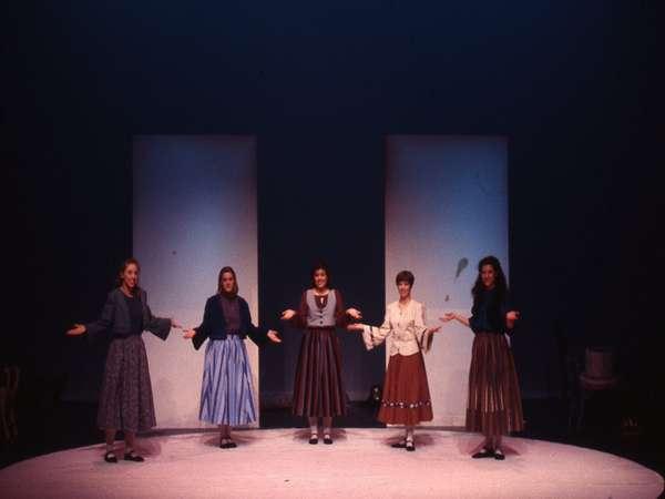 Five performers on stage, each wearing a long skirt and blouse, stand in evenly spaced positions with their arms outstretched to the sides. They are illuminated by soft lighting against a dark background with three tall panels behind them.