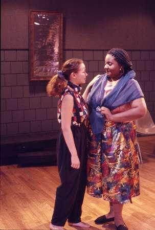 Two people face each other in a room with wooden floors and a gray brick wall. One wears a patterned shirt and dark pants, the other a colorful skirt with a blue shawl. A framed picture hangs on the wall behind them.