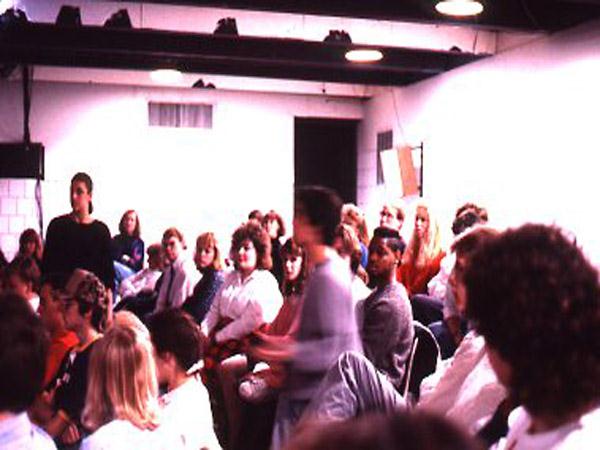 A group of people seated in a dimly lit room, engaged in what seems to be a discussion or seminar. One person stands while others sit attentively. The setting is informal with white walls and overhead lighting.