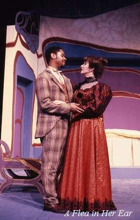A man and woman in period costumes stand facing each other on a colorful stage set. The woman wears a long red dress with a black lace top, and the man wears a plaid suit. The backdrop includes abstract shapes and patterns. Text reads, “A Flea in Her Ear.”.