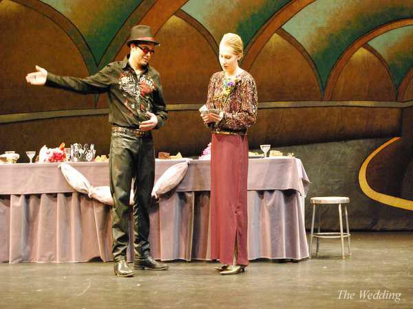 A man in a cowboy hat and embroidered shirt gestures while talking to a woman in a long skirt and patterned top. They stand in front of a table with a purple tablecloth on a stage with an ornate backdrop.