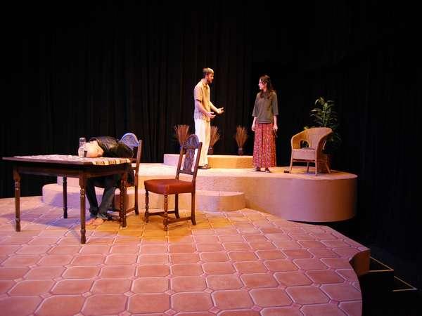 Three people on a stage: one sits slumped over a table, while two stand facing each other, gesturing expressively. The stage is set with a few chairs and plants, with a black curtain backdrop.