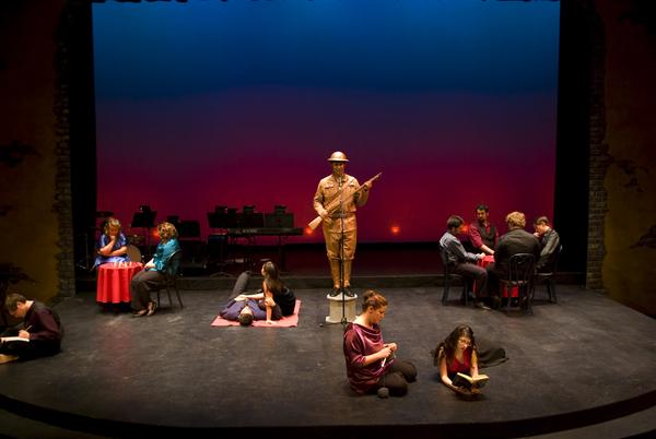 A theatrical stage scene with people lounging in pairs reading and talking. A soldier mannequin in uniform stands center stage holding a rifle. The background is lit in blue and pink, with musicians silhouetted in the back.