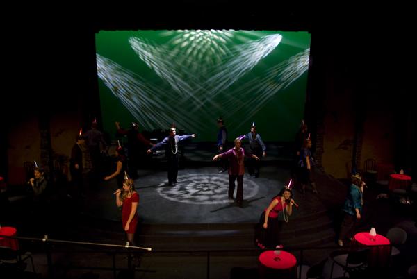 Performers on a stage with a green backdrop featuring crisscross light patterns. The group wears festive hats, and the scene includes red tabletops scattered around the stage, creating a celebratory atmosphere.
