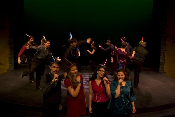 A group of people on stage wearing party hats and performing. Four individuals stand at the front with microphones, singing, while others dance energetically in the background. The stage is dimly lit, with a green backdrop.