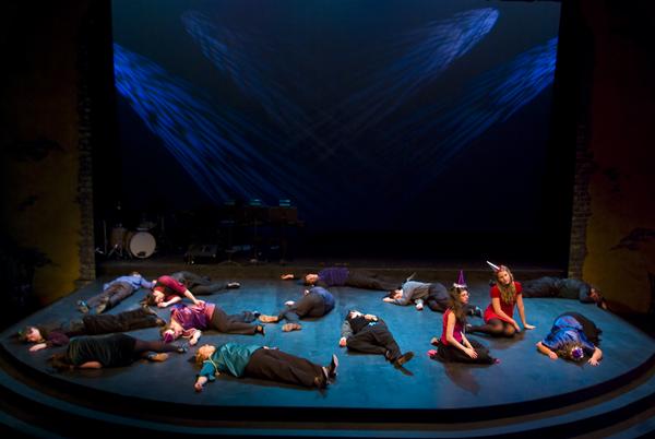A dimly lit stage scene with multiple performers lying scattered across the floor. A woman in a red dress is among them, sitting upright. The background features a piano and drums, with blue lighting casting a dramatic atmosphere.