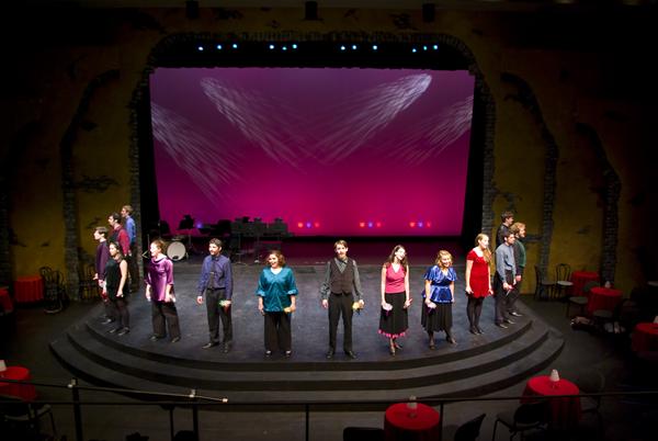 A group of performers stands on a theater stage with a pink and purple backdrop. They are dressed in various colorful outfits, standing in a semi-circle formation. The stage is set with minimal props and a lit background.