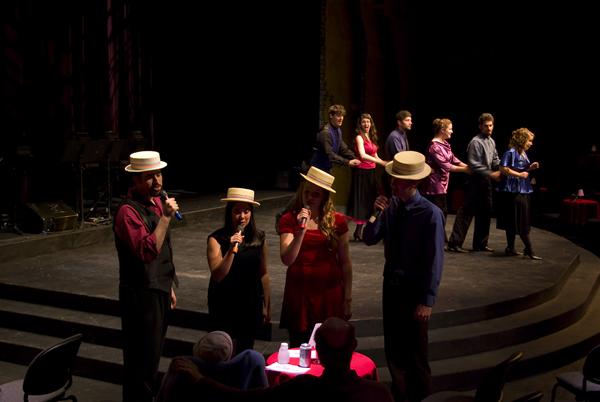 Four performers in straw hats sing into microphones on a dimly lit stage. They are dressed in vibrant attire. In the background, more performers stand in pairs. The stage is circular with audience members seated at small tables.