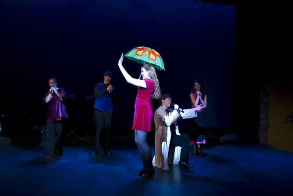 Performers on stage with dark background; one person holds a colorful umbrella. Another kneels, while others stand and smile, dressed in varying costumes. The scene is dramatic and likely part of a theatrical production.