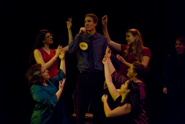 A male performer stands on stage holding a microphone, surrounded by five women reaching toward him. The group is engaged in a musical or theatrical performance with dramatic lighting.