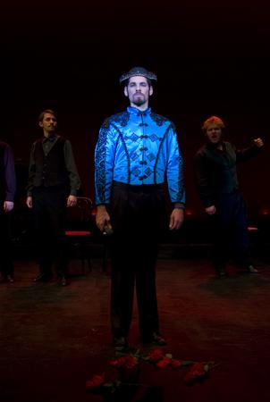 A man stands in the center of a dark stage wearing a shiny blue, patterned jacket and a black hat. Red roses are scattered at his feet. Two other men are visible in the background, blurred and wearing dark clothing.