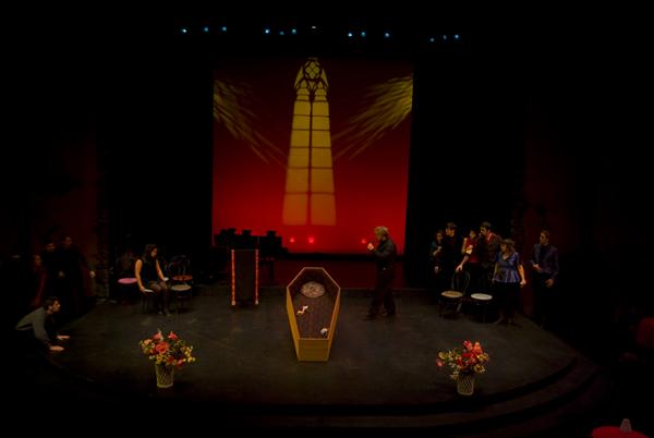A theater stage depicts a funeral scene with a central coffin and floral arrangements. Actors in mourning attire stand and sit around the stage. A large window projection with a red backdrop enhances the dramatic atmosphere.