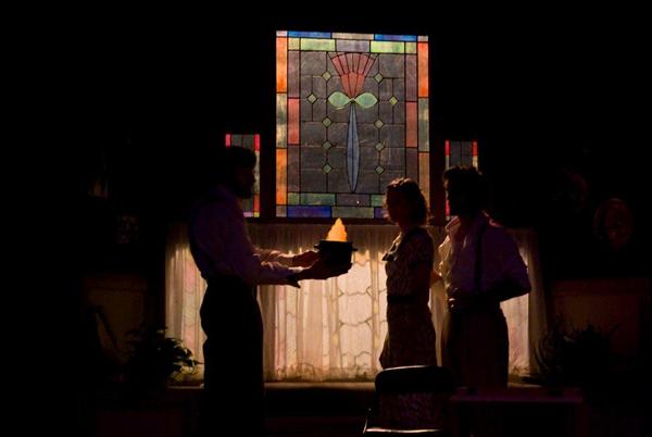 Three people in silhouette stand indoors near a table. One holds a lit object. Behind them is a colorful stained glass window depicting a flower. A bright light shines through a curtained window, casting dramatic shadows.