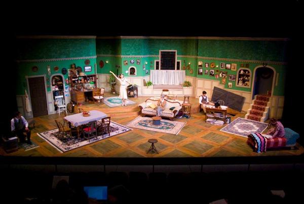 A theater stage set depicting a vintage living room with green wallpaper. The scene features a dining area, bookshelves, a sofa, a desk, and framed pictures on the walls. Five actors are seen performing various activities around the room.