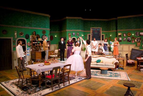 A staged play with actors in a room decorated with green walls and vintage furniture. Some actors are standing, while one in a white costume gestures animatedly. The scene includes a table, couch, and several framed pictures on the walls.