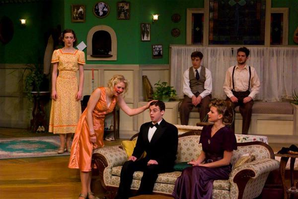 A theatrical scene with actors in vintage costumes. A woman in an orange dress leans toward a seated man in a tuxedo, while others watch from a sofa and standing positions. The set is designed to resemble a vintage living room.